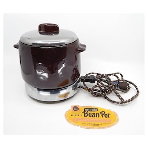 May include: A brown ceramic electric bean pot with a silver lid and base. The pot is on a white surface with a yellow and brown label that reads "West Bend Electric Bean Pot".