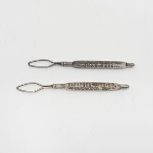 May include: Two vintage metal shoe horns with loop handles. One is marked "SWAN SHOE" and the other "TRUMBULL SHOE CO. WORCESTER, MASS.". The shoe horns are silver in color and appear to be made of metal.