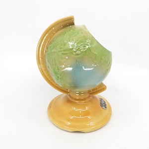 May include: A ceramic globe-shaped decorative item. The globe features a light green and blue color scheme, with a yellow stand and support. The item has a glossy finish and is likely a vintage or collectible piece.