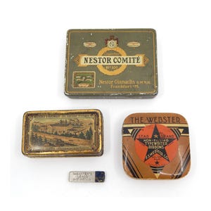 May include: An assortment of vintage metal containers, including a rectangular green and gold Nestor Comité cigarette tin, a rectangular tin with a landscape scene, and a square tin for typewriter ribbons. Also, a small metal Sheaffer's Leads container.