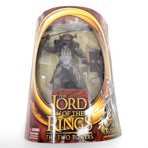 May include: A packaged action figure from "The Lord of the Rings: The Two Towers" featuring an Easterling warrior. The figure is dark with a spear and shield, displayed in a clear plastic window within a themed box.