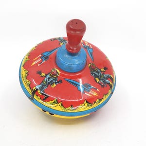 May include: Vintage metal spinning top with a red body featuring astronauts and rockets. The top has a blue base and a red handle. The design includes yellow and blue accents, creating a retro toy aesthetic.