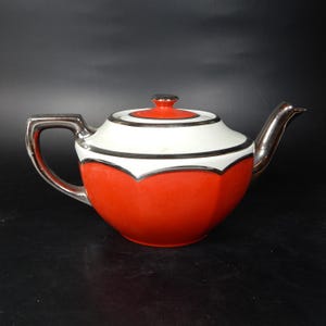 May include: A vintage teapot with a red base, white middle section, and silver accents. The lid is red with a silver top. The handle and spout are silver. The teapot has a geometric design.