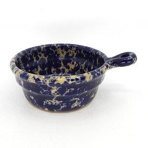 May include: A round ceramic bowl with a single handle on the side. The bowl features a dark blue base color with a speckled, cream-colored pattern throughout the interior and exterior surfaces, creating a mottled appearance on the glazed finish.