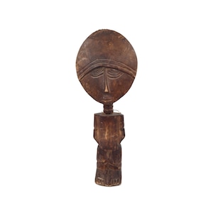 May include: A carved wooden sculpture featuring a stylized face with an oval head and detailed facial features. The sculpture is dark brown and stands on a carved base. The piece appears to be an African tribal art form.