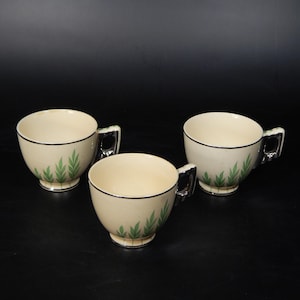 May include: Three cream-colored teacups with black trim and silver handles. Each cup features a green leaf design near the base. The cups are arranged on a black surface, showcasing a vintage tea set.