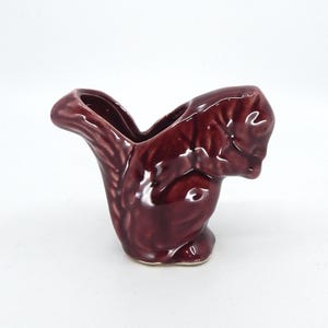 May include: A maroon ceramic squirrel-shaped creamer or small pitcher. The squirrel is depicted in a sitting position with its tail curved upwards, and the creamer has a small spout. The glossy finish enhances the details of the squirrel's fur.