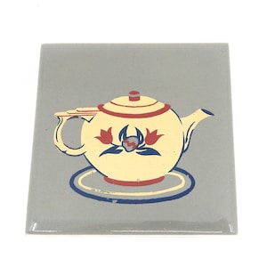 May include: A vintage ceramic tile with a gray background and a yellow teapot with red and blue floral designs. The teapot is sitting on a blue and white saucer.