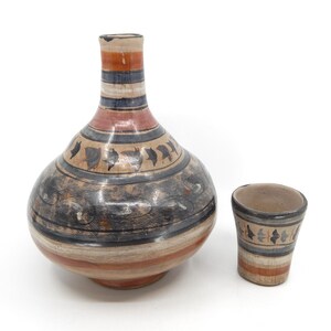 May include: A handcrafted ceramic water jug and cup set. The jug features a bulbous body and a long neck, adorned with horizontal bands of color and decorative patterns. The cup has a similar design. The color palette includes shades of brown, black, red, and cream.