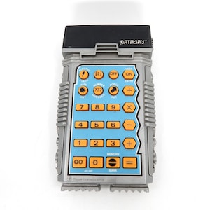 May include: A vintage Dataman calculator toy in silver and black. The calculator has a blue keypad with orange buttons and a variety of function keys. The text "DATAMAN" is printed at the top. The calculator is made by Texas Instruments.