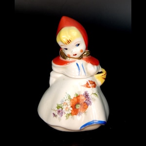 May include: A vintage ceramic sugar bowl shaped like a girl in a red hooded dress. The girl has blonde hair, blue eyes, and rosy cheeks. The dress is decorated with a floral pattern in shades of orange, purple, and green. The bowl has a yellow lid with a basket handle.