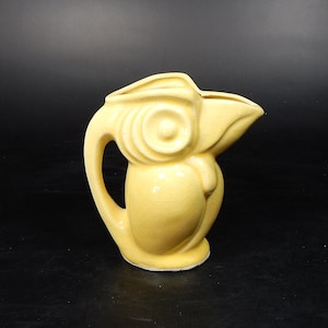 May include: A vintage, pale yellow ceramic pitcher with a unique owl-like design. The pitcher features a curved handle, a spout, and a textured surface. The owl's face is depicted with concentric circles. The pitcher is approximately 6 inches tall.