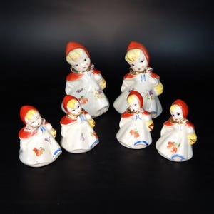 May include: Set of six ceramic figurines of Little Red Riding Hood. The figurines are all wearing red hoods and white dresses with floral patterns. They are holding baskets and are in various poses.
