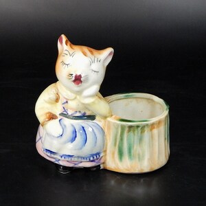 May include: A ceramic planter shaped like a cat wearing a dress, with closed eyes and a relaxed pose. The cat is white with brown stripes and a red mouth. The planter is a light beige with green and yellow accents.