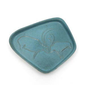 May include: A turquoise ceramic tray with a raised, etched design of two animals facing each other. The tray has a slightly irregular, rectangular shape with rounded corners. The animals have large horns.