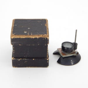 May include: A vintage black box with a worn, aged appearance sits next to a black magnifying glass with a clear lens. The magnifying glass is mounted on a black base with a metal rod.