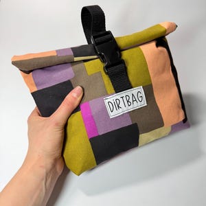 May include: A colorful, patchwork-style lunch bag with a black buckle closure and strap. The bag features a variety of colors, including orange, purple, green, and black. The text "DIRTBAG" is printed on a white label.