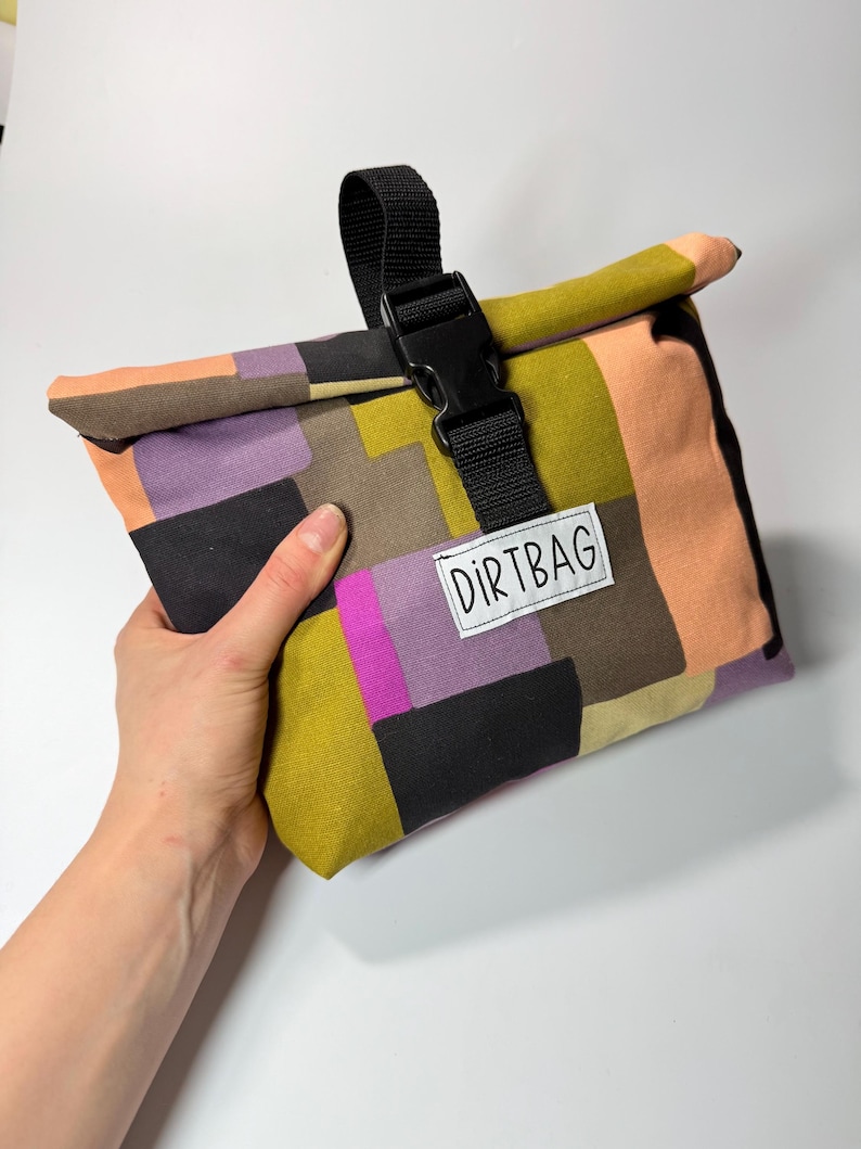 May include: A colorful, patchwork-style lunch bag with a black buckle closure and strap. The bag features a variety of colors, including orange, purple, green, and black. The text "DIRTBAG" is printed on a white label.