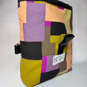 May include: A colorful chalk bag with a geometric pattern in shades of purple, orange, green, and black. The bag has a black top and a black buckle closure. The word "DIRTBAG" is printed on a white label.