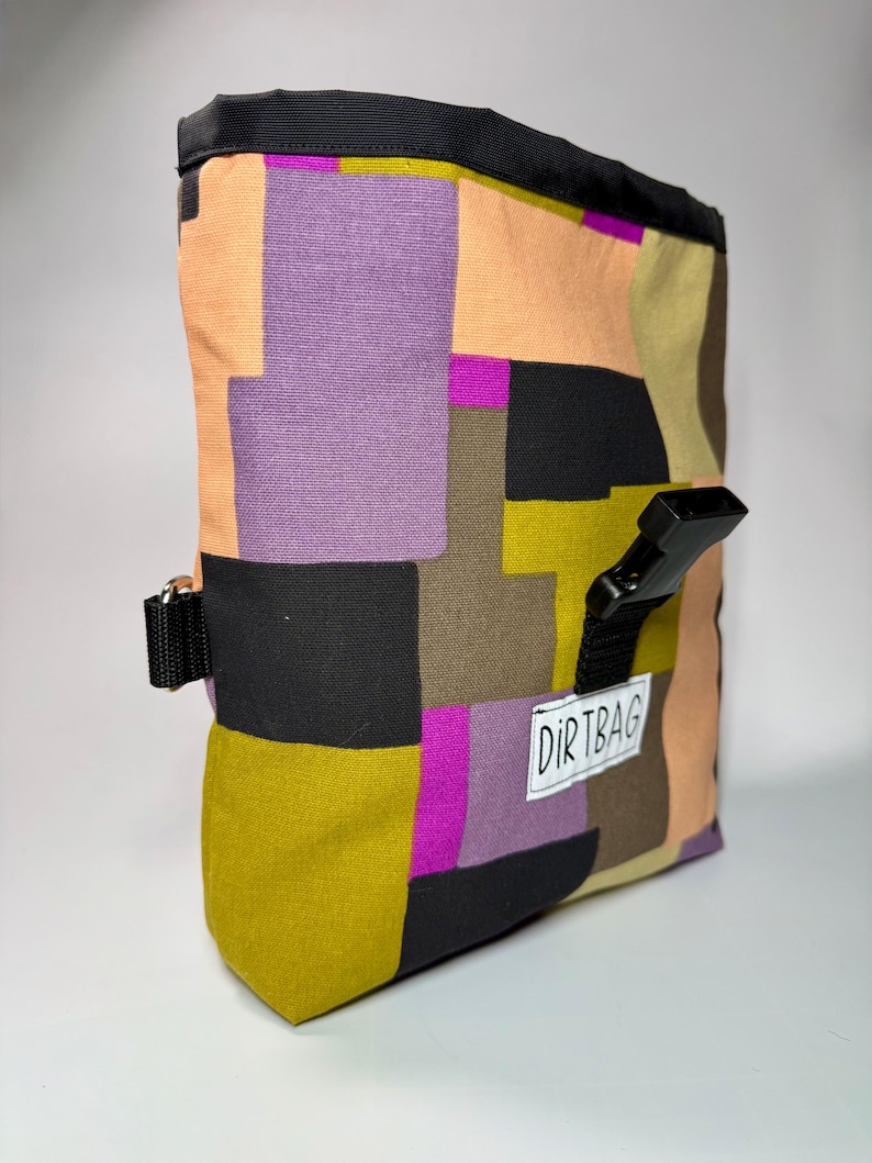 May include: A colorful chalk bag with a geometric pattern in shades of purple, orange, green, and black. The bag has a black top and a black buckle closure. The word "DIRTBAG" is printed on a white label.