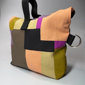 May include: A colorful patchwork bag with a black handle and a silver metal ring. The bag features a variety of colors, including orange, purple, green, and black, arranged in a geometric pattern. The bag is designed for carrying items.