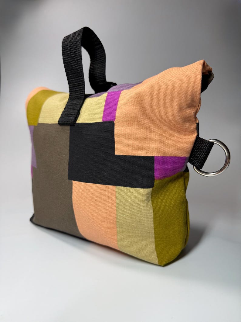May include: A colorful patchwork bag with a black handle and a silver metal ring. The bag features a variety of colors, including orange, purple, green, and black, arranged in a geometric pattern. The bag is designed for carrying items.