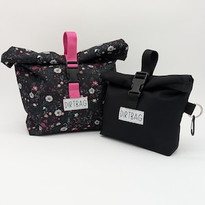May include: Two black fabric bags with a roll-top closure. The larger bag has a floral print and a pink strap with a buckle. The smaller bag has a black strap with a buckle and a key ring. Both bags have a white label with the text "DIRTBAG" printed on them.