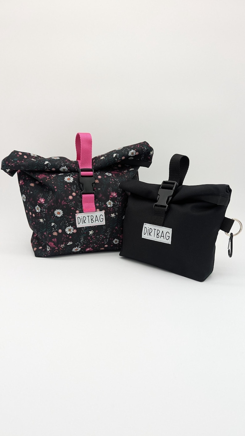 May include: Two black fabric bags with a roll-top closure. The larger bag has a floral print and a pink strap with a buckle. The smaller bag has a black strap with a buckle and a key ring. Both bags have a white label with the text "DIRTBAG" printed on them.