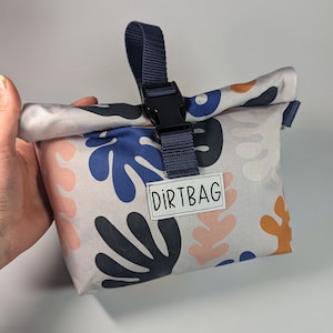 Może przedstawiać: Mała, szara torba z tkaniny z niebieskim paskiem i czarną klamrą. Torba ma białą etykietę z napisem "DIRTBAG". Torba jest ozdobiona wzorem abstrakcyjnych kształt&oacute;w w kolorze niebieskim, r&oacute;żowym i pomarańczowym.