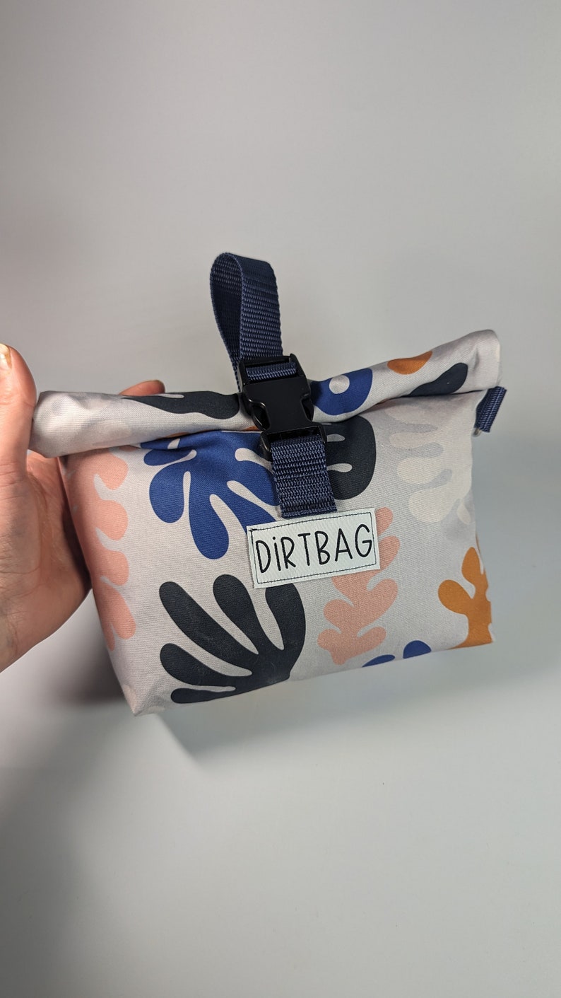 Może przedstawiać: Mała, szara torba z tkaniny z niebieskim paskiem i czarną klamrą. Torba ma białą etykietę z napisem "DIRTBAG". Torba jest ozdobiona wzorem abstrakcyjnych kształt&oacute;w w kolorze niebieskim, r&oacute;żowym i pomarańczowym.