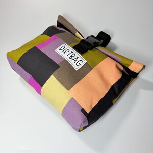 May include: A colorful patchwork bag with a black buckle closure. The bag features a variety of colors, including olive green, black, peach, purple, and yellow. The text "DIRTBAG" is printed on a white label on the front of the bag.