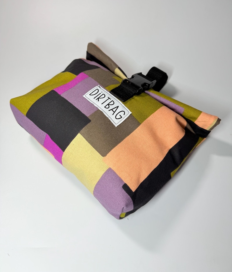 May include: A colorful patchwork bag with a black buckle closure. The bag features a variety of colors, including olive green, black, peach, purple, and yellow. The text "DIRTBAG" is printed on a white label on the front of the bag.