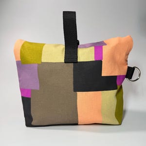 May include: A colorful patchwork bag with a black handle and a silver metal ring. The bag features squares of olive green, peach, purple, black, and tan fabric. The bag is designed to be a doorstop.