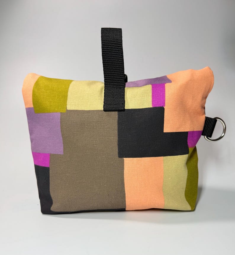 May include: A colorful patchwork bag with a black handle and a silver metal ring. The bag features squares of olive green, peach, purple, black, and tan fabric. The bag is designed to be a doorstop.