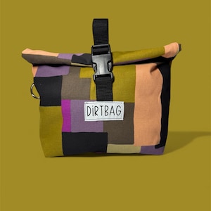 May include: A colorful patchwork bag with a black buckle closure and a black strap. The bag features a variety of colors, including black, purple, olive green, and peach. The word "DIRTBAG" is printed on a white label.