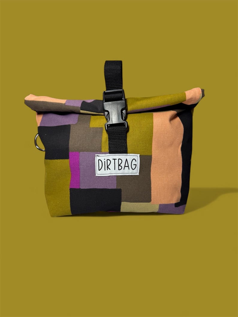 May include: A colorful patchwork bag with a black buckle closure and a black strap. The bag features a variety of colors, including black, purple, olive green, and peach. The word "DIRTBAG" is printed on a white label.