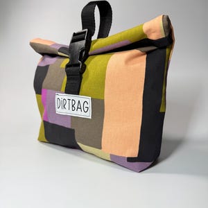 May include: A colorful roll-top bag with a patchwork design. The bag features a black buckle closure and a black handle. The text "DIRTBAG" is printed on a white label. The bag's color palette includes shades of green, orange, black, purple, and brown.