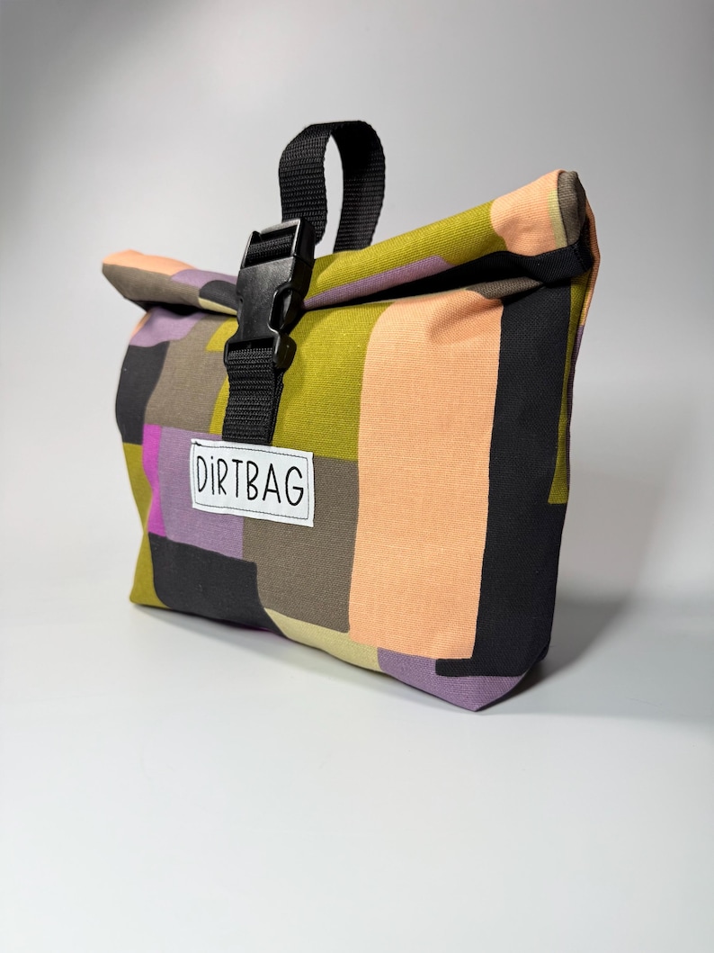 May include: A colorful roll-top bag with a patchwork design. The bag features a black buckle closure and a black handle. The text "DIRTBAG" is printed on a white label. The bag's color palette includes shades of green, orange, black, purple, and brown.