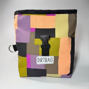 May include: A colorful, patchwork-style chalk bag with a black top and buckle closure. The bag features a mix of purple, orange, green, and black fabric squares. A white label reads "DIRTBAG". A metal ring is attached to the side.