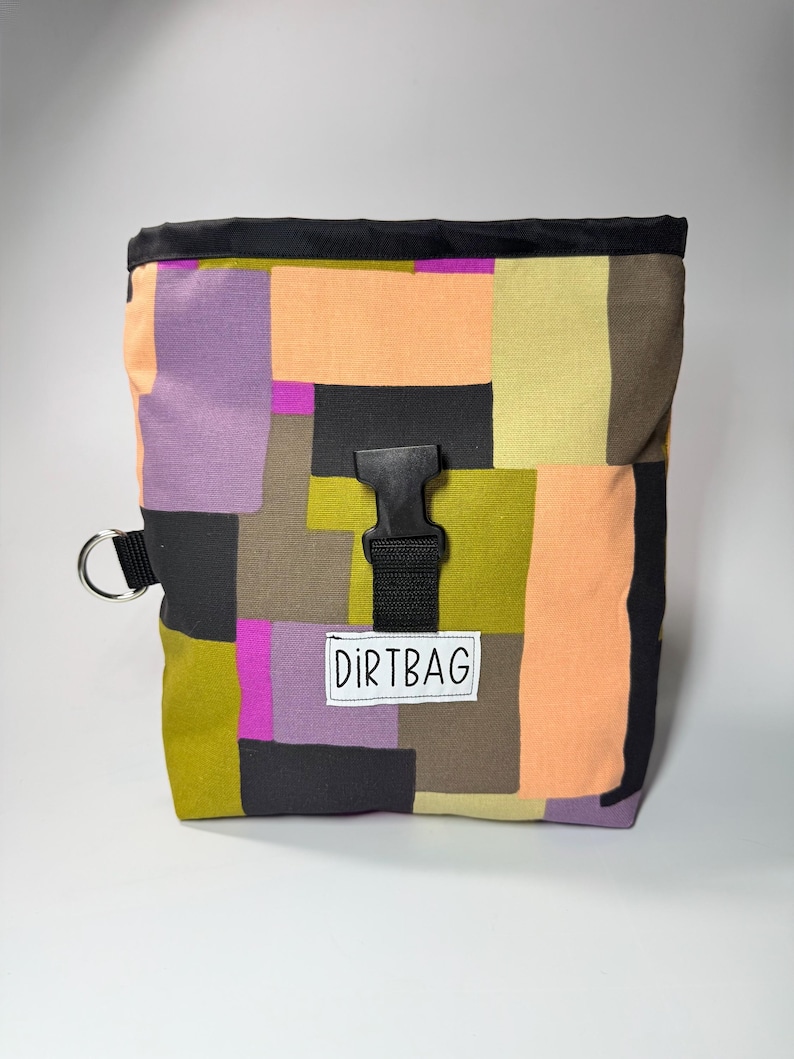May include: A colorful, patchwork-style chalk bag with a black top and buckle closure. The bag features a mix of purple, orange, green, and black fabric squares. A white label reads "DIRTBAG". A metal ring is attached to the side.