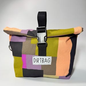 May include: A colorful roll-top bag with a patchwork design in shades of green, purple, orange, and black. The bag features a black buckle closure and a small white label that reads "DIRTBAG".