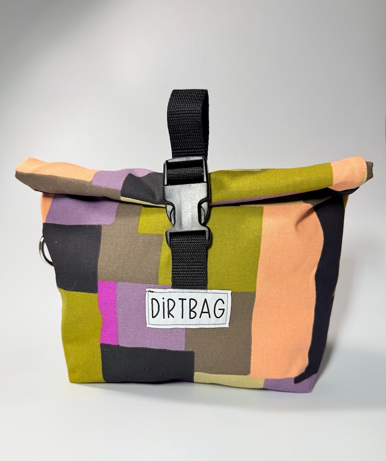 May include: A colorful roll-top bag with a patchwork design in shades of green, purple, orange, and black. The bag features a black buckle closure and a small white label that reads "DIRTBAG".