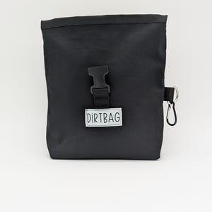 May include: A black canvas bag with a black buckle and a silver clip. The bag has a white label that says "DIRTBAG".