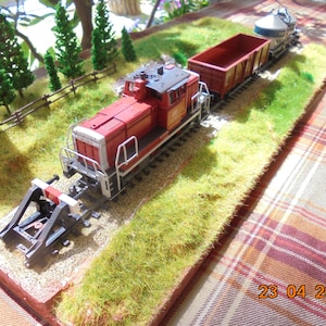 May include: A model train set featuring a red and silver diesel locomotive pulling two freight cars on a miniature track. The scene includes miniature trees, a fence, and a grassy area, all set on a brown base.