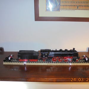 May include: A black model train with red wheels and a black tender sits on a track. The train is on a wooden surface. The date "24 03 2026" is visible on the surface.