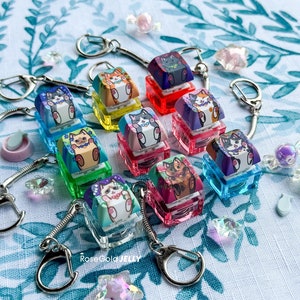 Puede incluir: Una colección de diez llaveros coloridos con diferentes diseños de gatos. Cada llavero está hecho de plástico transparente con una base colorida y una imagen impresa de un gato. Los llaveros están unidos a llaveros de plata.