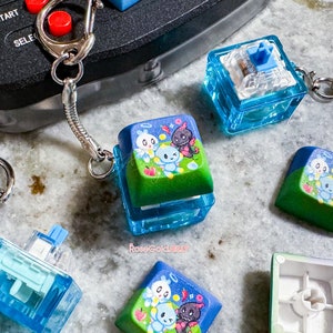 May include: A collection of custom keycaps with a cartoon design. The keycaps feature a blue and green color scheme with a cartoon character design. Some keycaps are attached to a keychain.