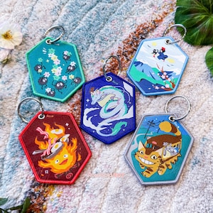 May include: Five colourful keychains featuring characters from popular animated films. The keychains are hexagonal shaped and feature embroidered designs. The keychains are on a white fluffy surface.