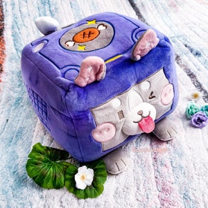 Puede incluir: Un juguete de peluche con forma de cubo con la cara de un perro en un lado. El cubo es morado con una cara gris y orejas rosas. El perro tiene una cara blanca con una nariz rosa y la lengua fuera. El juguete está sobre una superficie peluda blanca y azul.