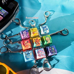 May include: A set of nine colorful keychains featuring different Pokemon characters. Each keychain is a square, clear acrylic charm with a different Pokemon character printed on it. The keychains are attached to silver chains with silver key rings.